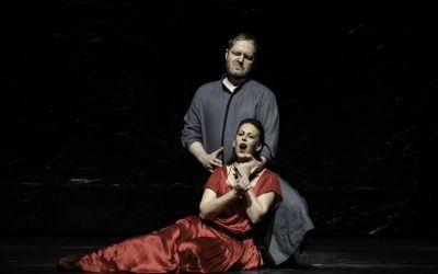 na scenie siedzi śpiewająca kobieta, nad nią klęczy mężczyzna ubrany w szarą szatę, przytrzymuje kobietę za szyje, czarne tło