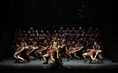 scena grupowa na froncie tancerze ubrani w czerwień tańczą w parach, kobiety leżą, mężczyźni je przytrzymują, w tle siedzą osoby w karmazynowych strojach, czarne tło