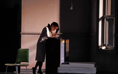 na ciemnym tle stoi zmartwiona kobieta opierająca się o czarną skrzynię, po jej prawej stronie łóżko, po lewej zielone krzesło, z góry zwisa zgaszona żarówka, po prawej okno oraz telefon